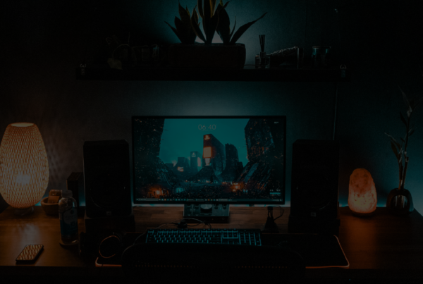 Scrivania su cui è poggiato un monitor pc e una tastiera retroilluminata in condizioni di scasa illuminazione. Sono presenti degli elementi decorativi su come delle lampade e delle piante. Dietro la scrivania è presente una strisce led lungo tutta la sua larghezza.