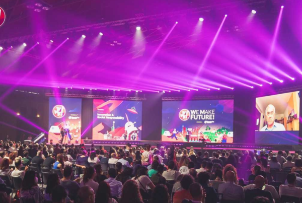 Una grande conferenza con un pubblico seduto, illuminazione colorata sul palco, schermi multipli con la scritta “We Make Future” e un relatore in videochiamata.