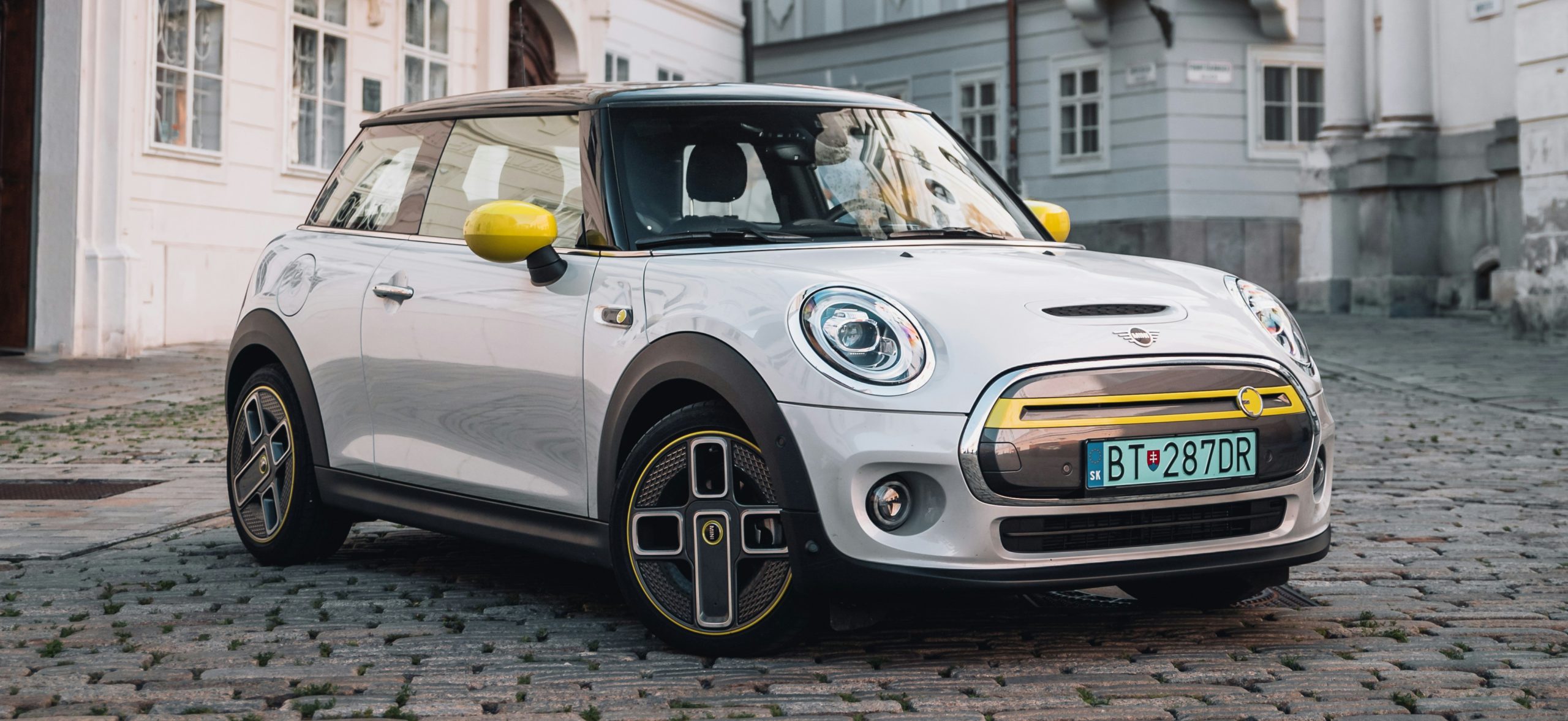 Mini Cooper SE , grigia con specchietti retrovisori gialli parcheggiata in modo inclinato verso destra, in un paese.