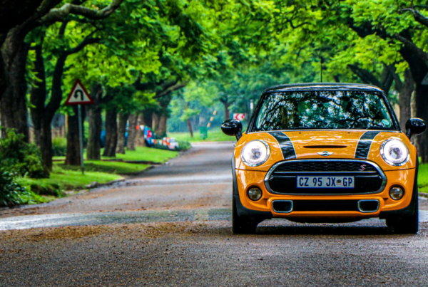 Yellow Mini Cooper S with black stripes walking on a tree-lined street in a residential area on a cloudy day.